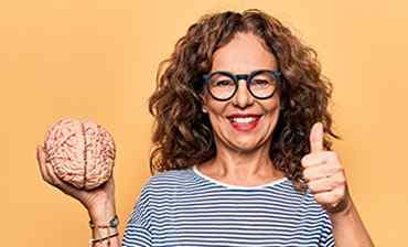 a mature woman smiling with fake brain in one hand and doing thumbs up from the other hand