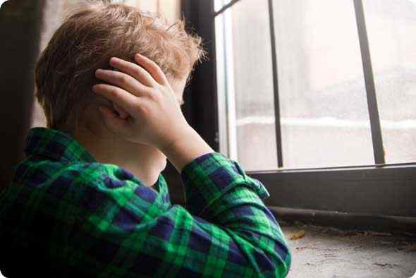 a male autistic child looking across the window covering his ears wth his hands